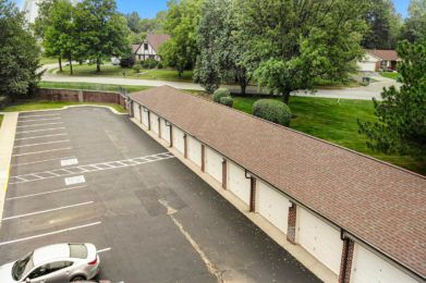 A row of storage units next to a parking lot surrounded by greenery and a nearby road.