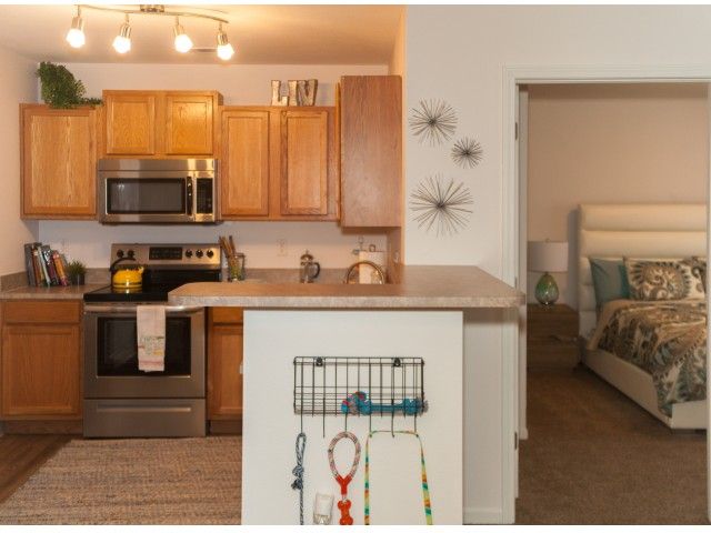 A modern apartment kitchen with wooden cabinets and stainless steel appliances, opening into a cozy living area.