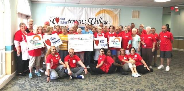 A group of people gathered indoors for a community event, featuring a large check presentation and participants wearing matching T-shirts.