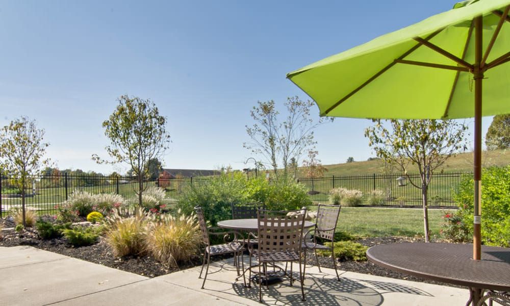 A sunny patio area with a green umbrella, table, and chairs set amidst a landscaped garden.