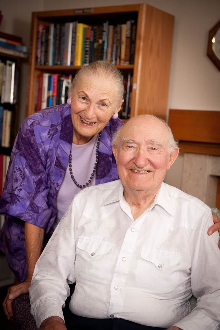 An elderly couple smiling together in a room with a bookshelf in the background.