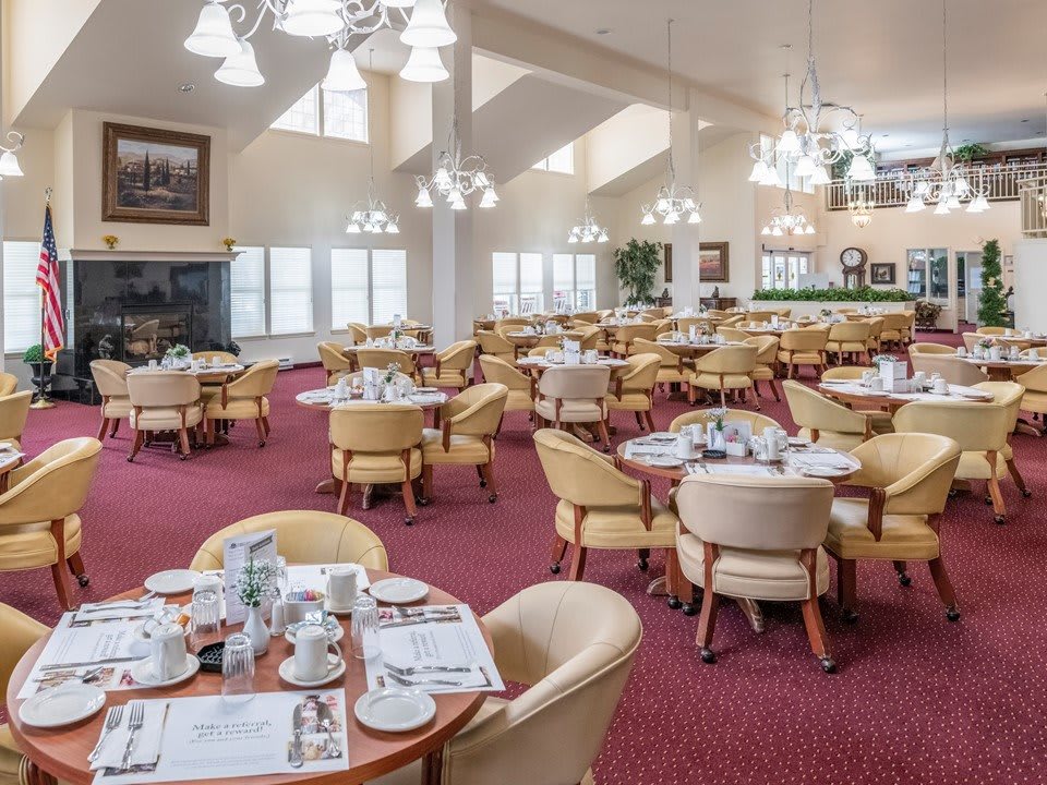 A spacious restaurant dining room with elegant chandeliers and neatly arranged tables and chairs on red carpet flooring.
