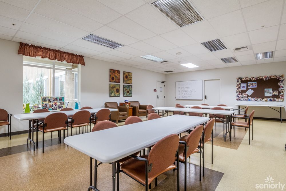A classroom or meeting room interior with tables and chairs arranged in rows, featuring a whiteboard and large windows.
