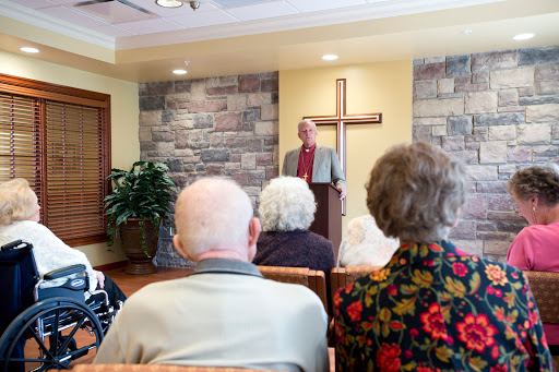A speaker addresses a group of elderly attendees in a church setting, with a cross prominently displayed in the background.