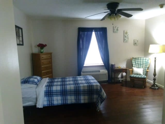 A cozy bedroom with a single bed and blue plaid bedding, featuring a dresser, chair, and window with blue curtains.