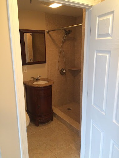 A view of a small bathroom with a shower, sink, and mirror through an open doorway.