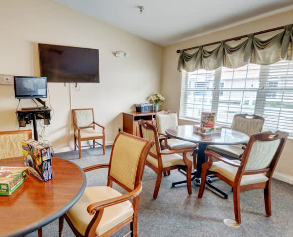 A cozy seating area with tables, chairs, a television, and a large window with curtains.