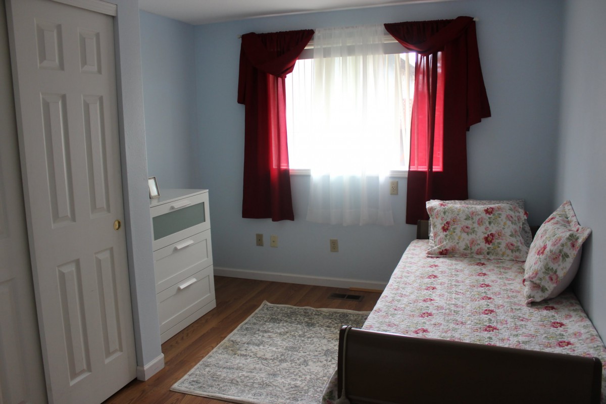 A cozy bedroom featuring a single bed with floral bedding, a white dresser, and red curtains over a window.