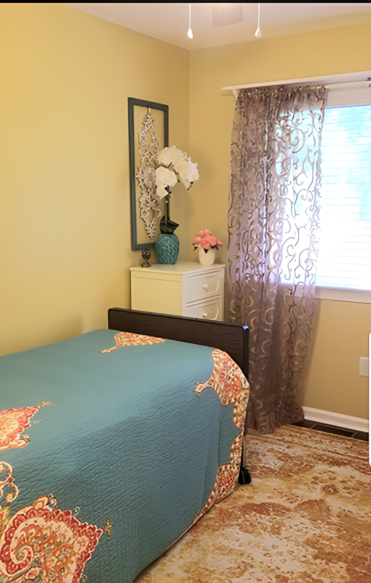 A cozy bedroom featuring a bed with a colorful quilt, decorative curtains, and a vase with flowers on the dresser.