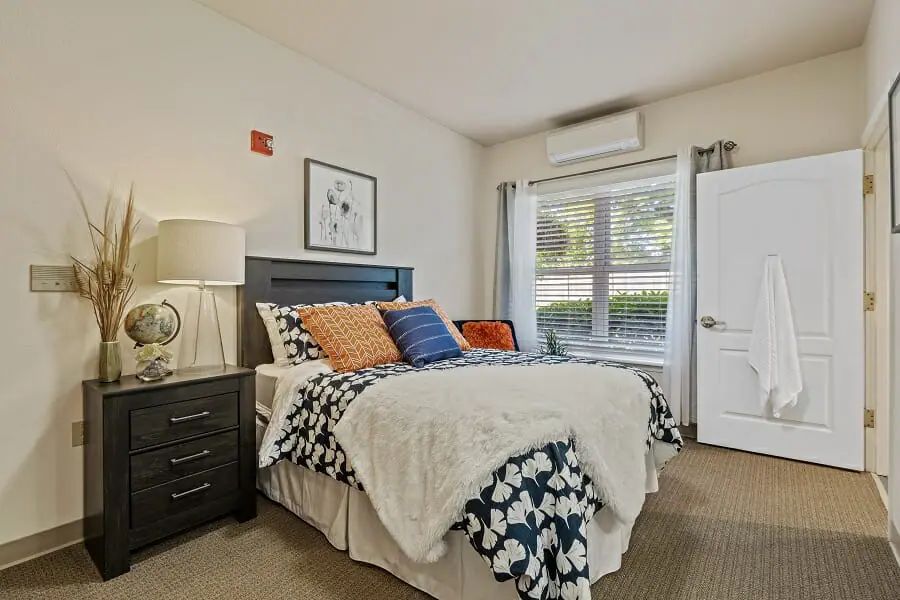 A cozy bedroom with a bed adorned with pillows and a bedside table, illuminated by natural light from a large window.
