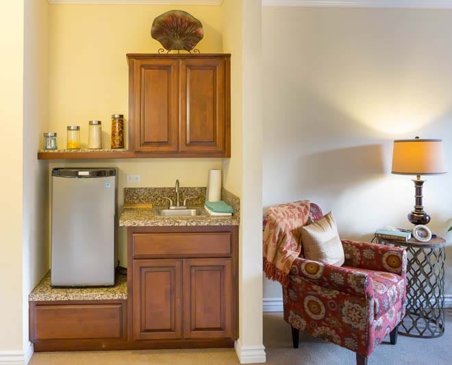 A cozy living space featuring a kitchenette with cabinets, a sink, and a mini fridge adjacent to an armchair and lamp.