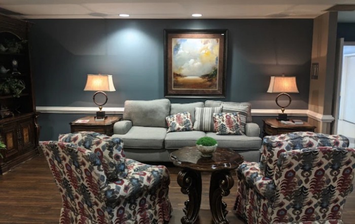 A cozy living room setting featuring a sofa with decorative cushions, two patterned armchairs, and a central coffee table. Two lamps provide lighting on either side of the sofa, with a landscape painting on the wall.