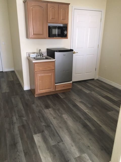 A small kitchenette area with wooden flooring, featuring a mini fridge, microwave, sink, and cabinetry.