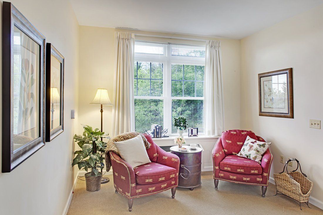 A cozy living room corner with two patterned armchairs, a side table, a floor lamp, and a large window providing natural light.
