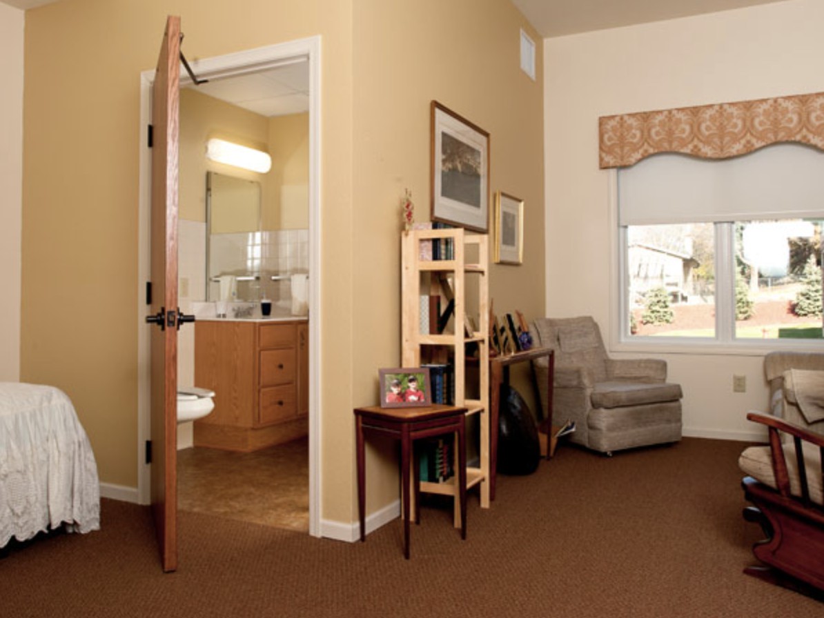 A cozy living room with a chair, bookshelf, and an open door leading to a bathroom.