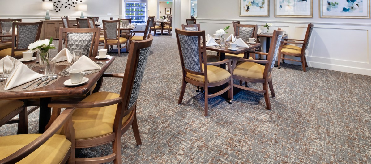 A well-organized dining room with wooden chairs and tables set for a meal, featuring a patterned carpet and bright lighting.