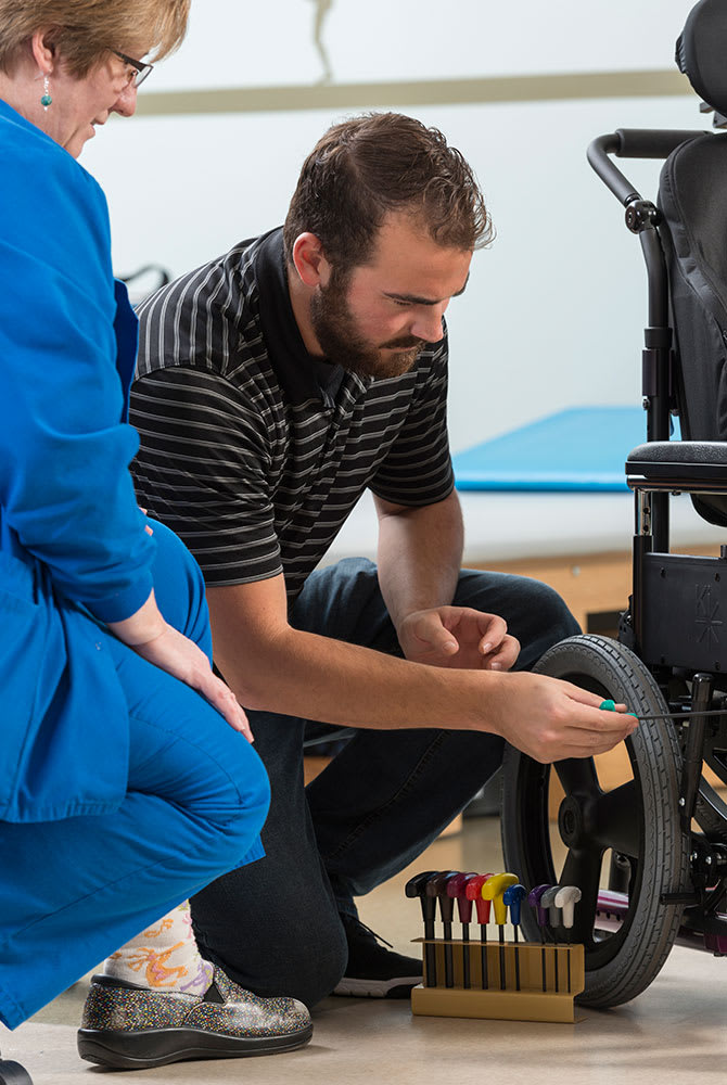 A professional performs maintenance on a wheelchair while discussing with a colleague.