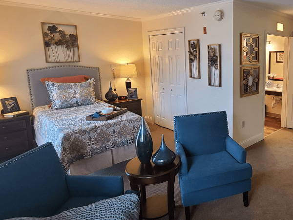 A cozy bedroom with a patterned bedspread, two blue chairs, and tasteful artwork on the walls.