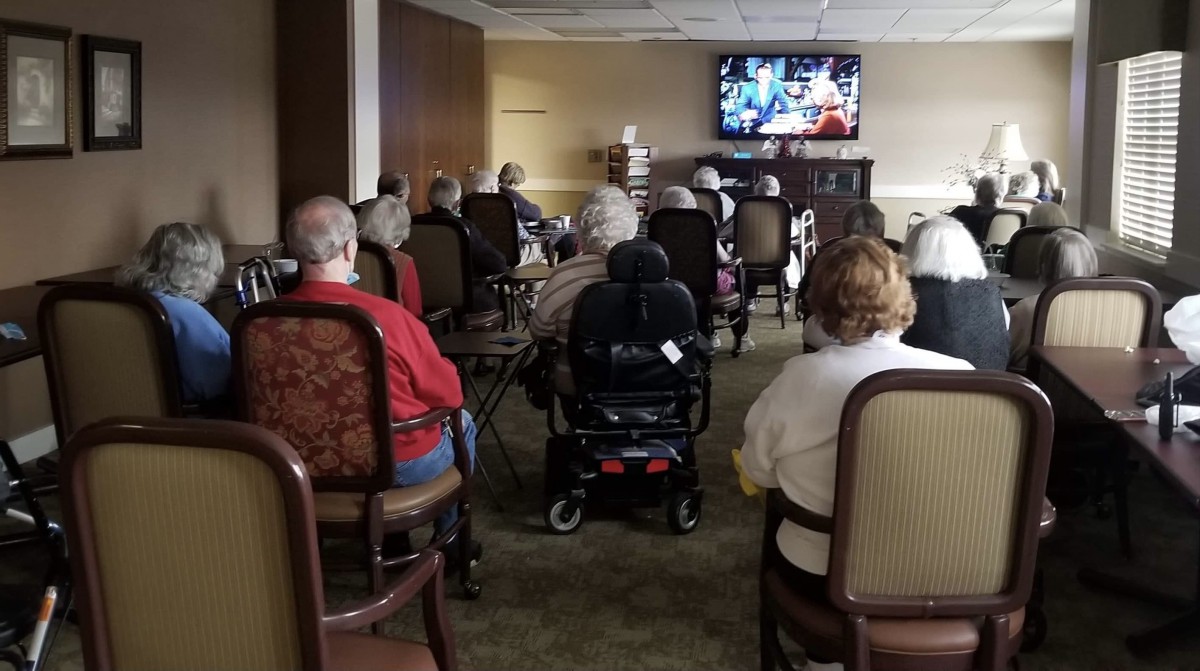 A group of elderly people watching television in a community room, with one person in a wheelchair.