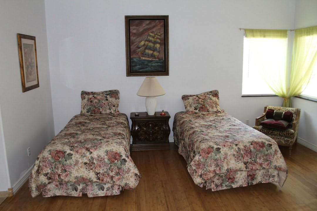 A cozy bedroom with two twin beds featuring floral bedding, separated by a nightstand with a lamp and adorned with a ship painting on the wall.