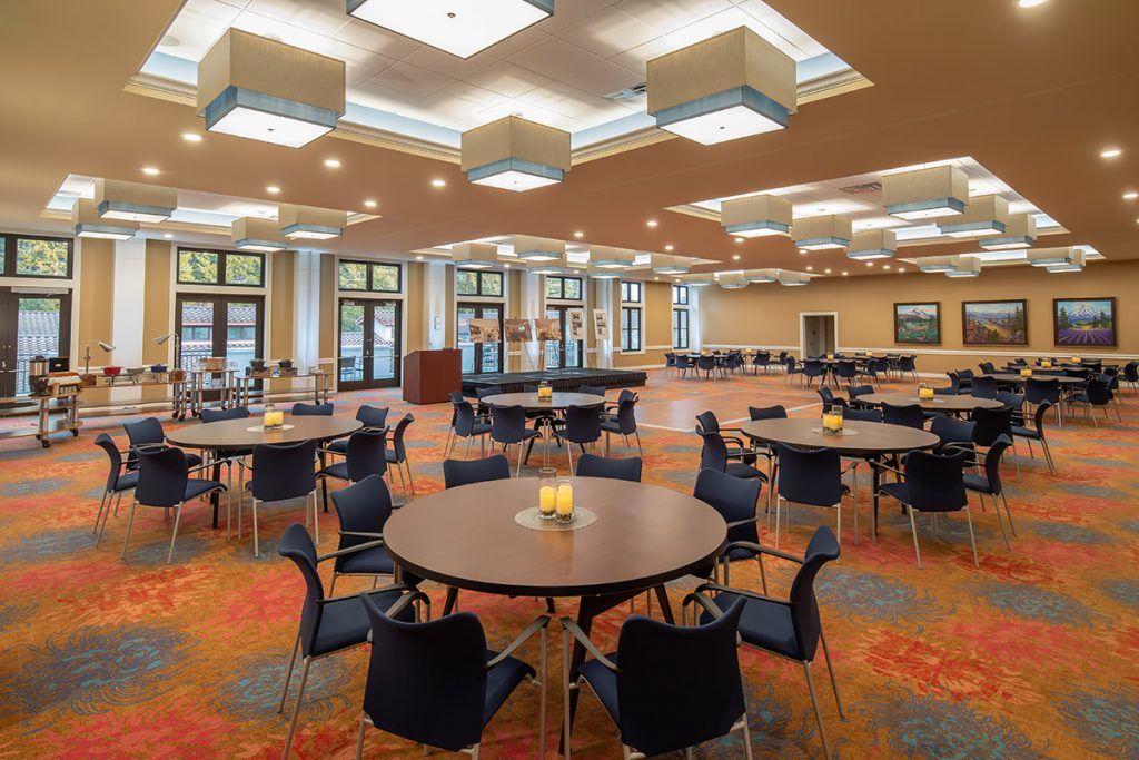 A spacious banquet hall with round tables and chairs arranged for an event, featuring modern lighting fixtures and large windows.
