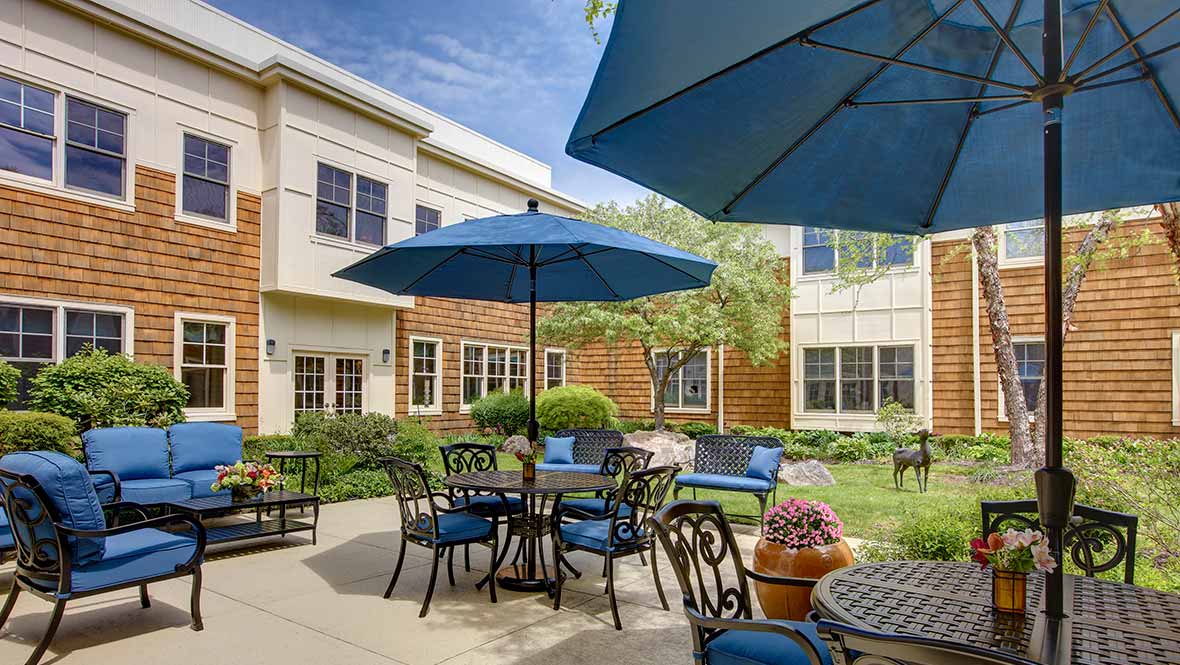 A sunny outdoor patio area with tables, chairs, and large blue umbrellas next to a building with well-maintained landscaping.
