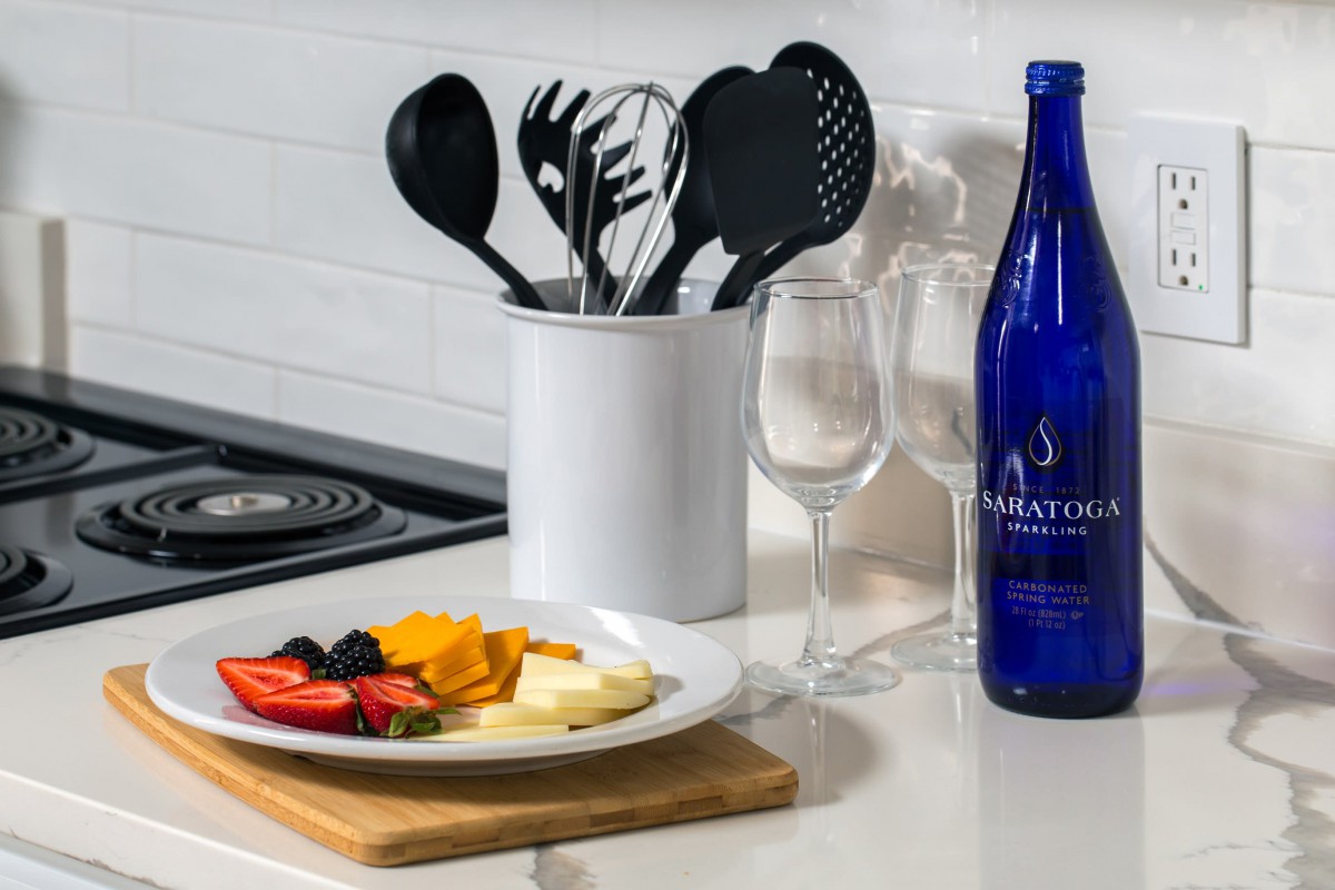 A kitchen countertop with utensils in a holder, a fruit platter, and a blue water bottle beside wine glasses.