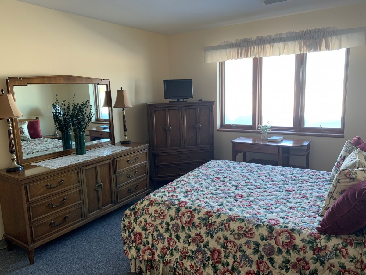 A cozy bedroom featuring a floral bedspread, wooden furniture, and a sunny window view.