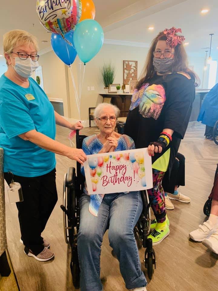 A joyful birthday celebration with a group of individuals, including an elderly person in a wheelchair holding a 'Happy Birthday' sign, surrounded by balloons.