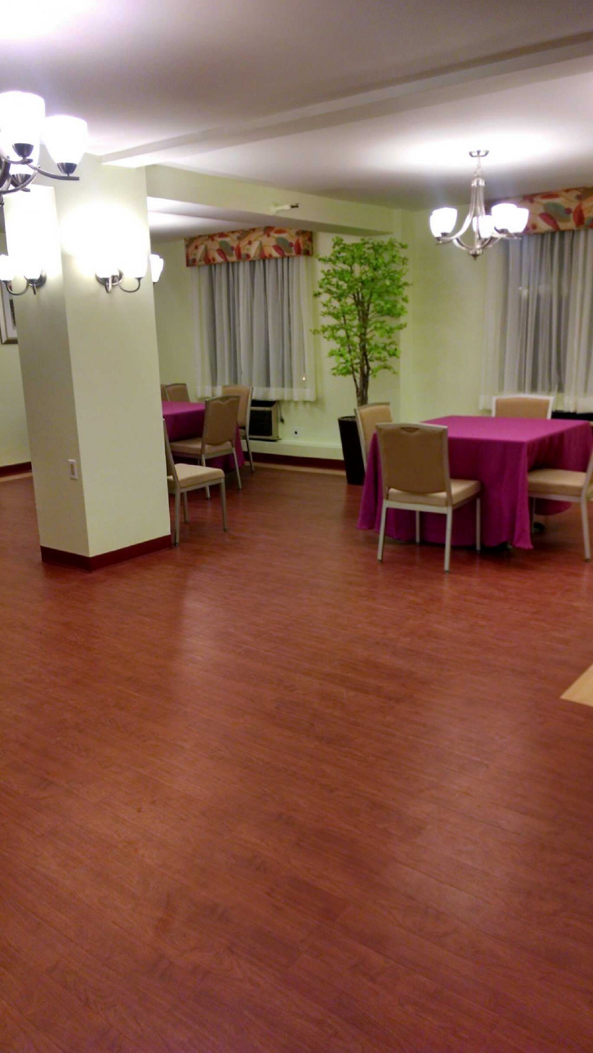 An empty conference or dining room with tables and chairs arranged around the space, featuring a wooden floor and decorative plants.