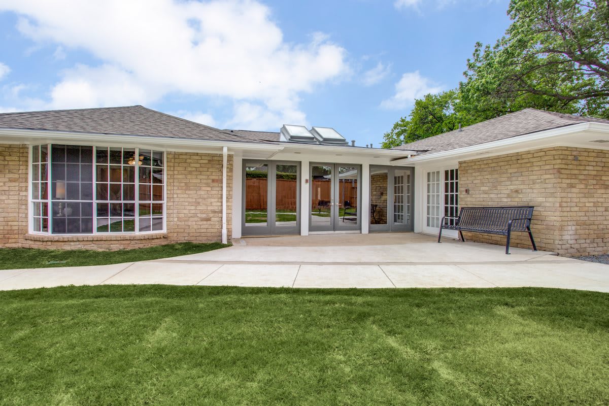 A single-story house with a spacious backyard and patio area, featuring large windows and a well-maintained lawn under a partly cloudy sky.