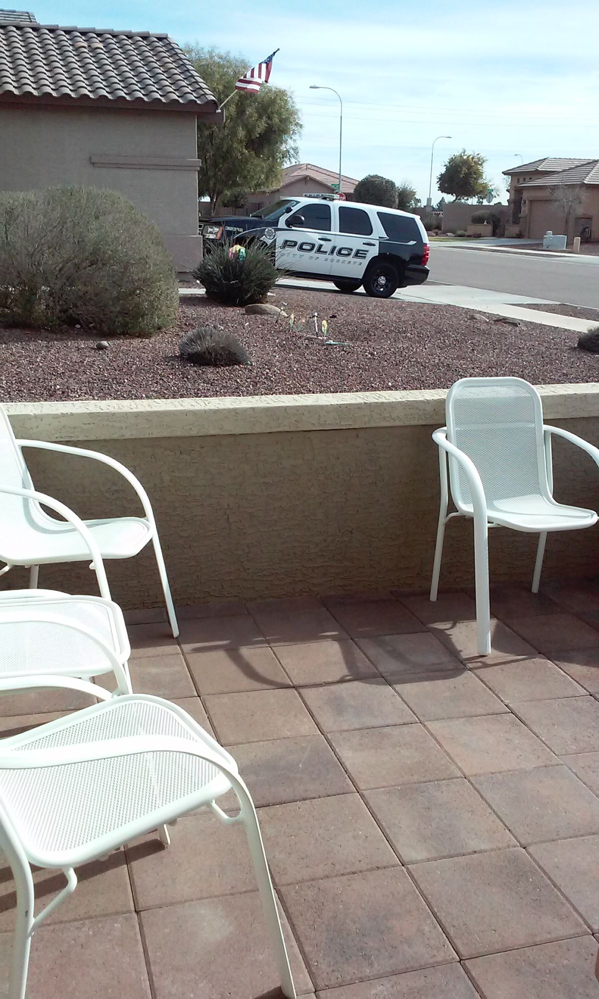 A residential patio with white chairs overlooks a driveway with a parked police car.