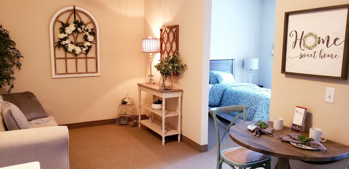 A cozy interior featuring a bedroom with blue bedding, a side table with a lamp and plants, and warm-toned walls.