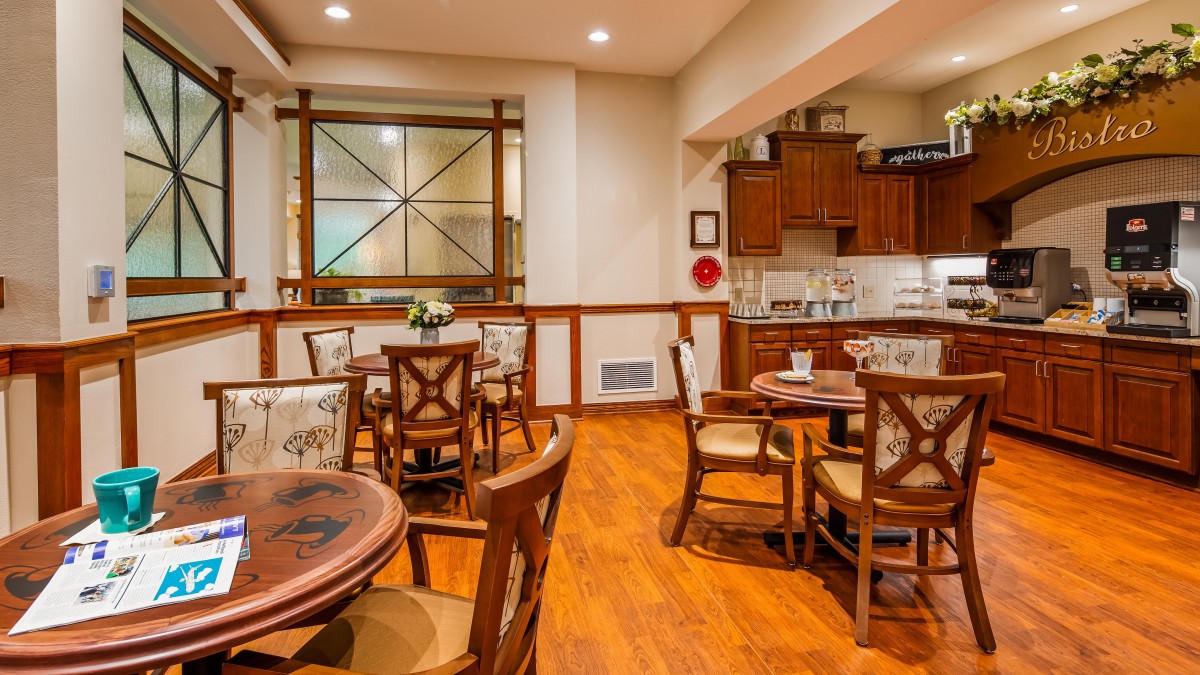 A cozy dining area with wooden tables and chairs, adjacent to a well-lit kitchen.