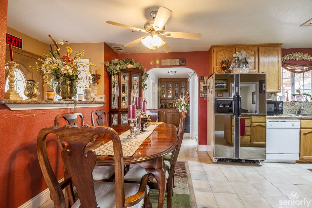 A cozy dining room with a traditional design, featuring a wooden table and chairs, ceiling fan, vibrant decor, and a view into the kitchen with stainless steel appliances.