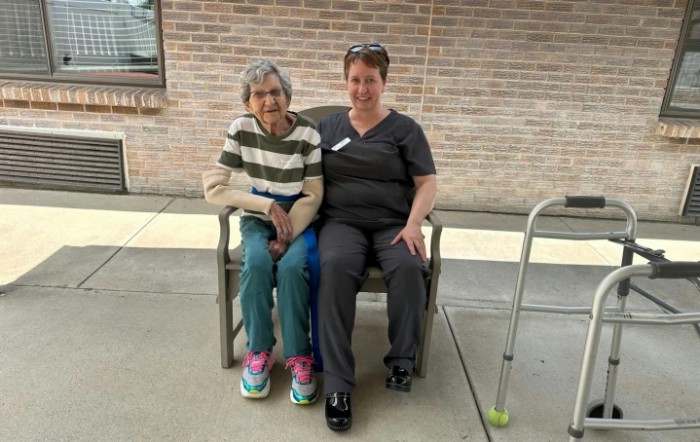 An elderly woman and a caregiver sitting together on a bench outside.