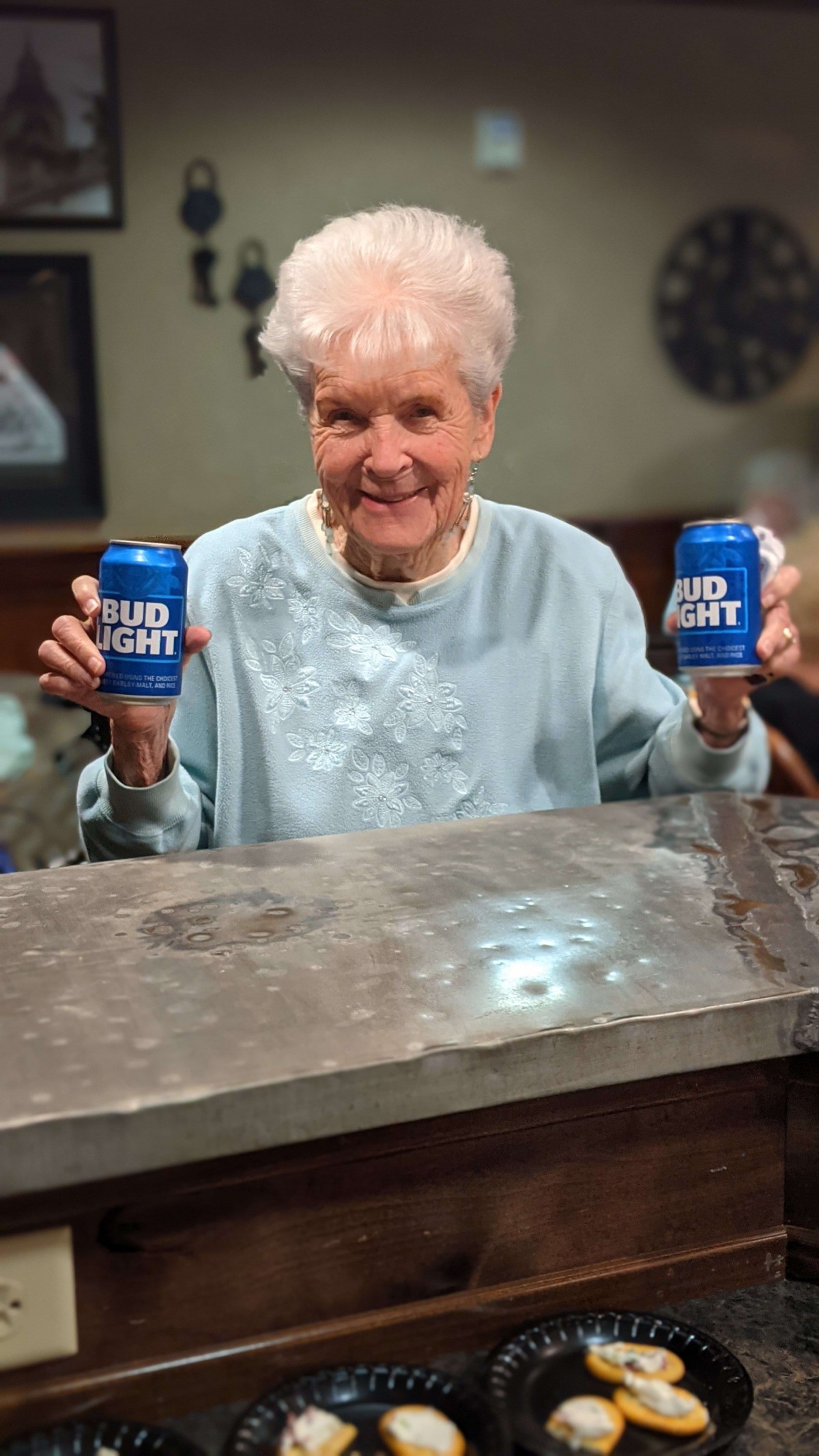 An elderly person smiling while holding two Bud Light cans indoors.