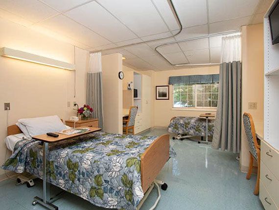 A well-furnished hospital room with two beds, floral bedspreads, a window, and privacy curtains.