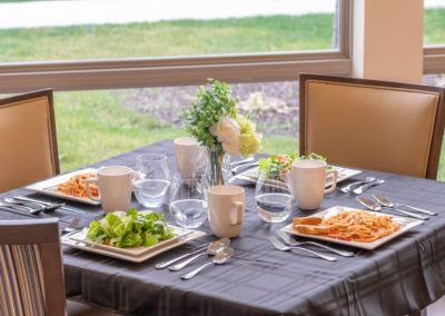 A neatly set dining table with plates of food and cutlery, positioned by a window providing a view of the outdoors.