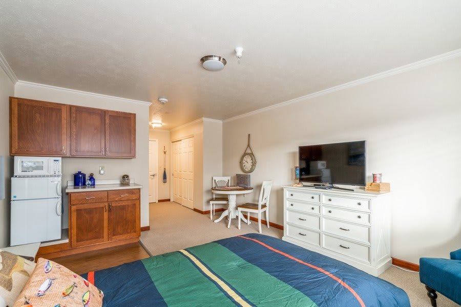 A cozy studio apartment with a kitchenette and modern furnishings, including a bed, dresser with TV, and small dining area.