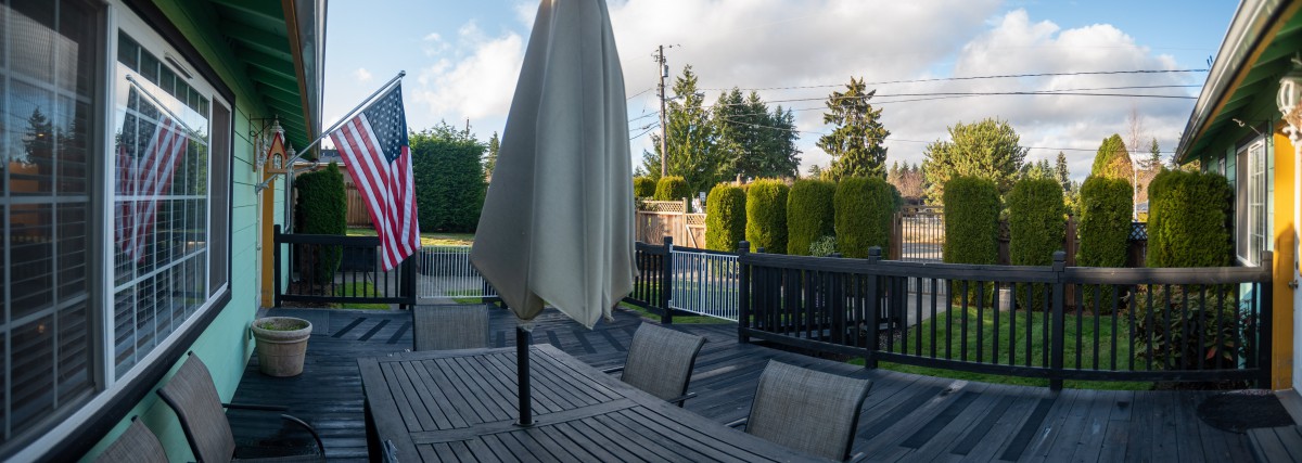 A tranquil patio setting with a table, chairs, and closed umbrella overlooking a fenced yard and trees.