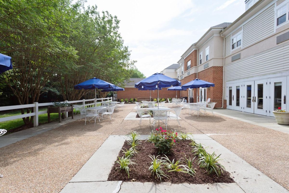 An outdoor patio area with tables and blue umbrellas, surrounded by greenery and a building.