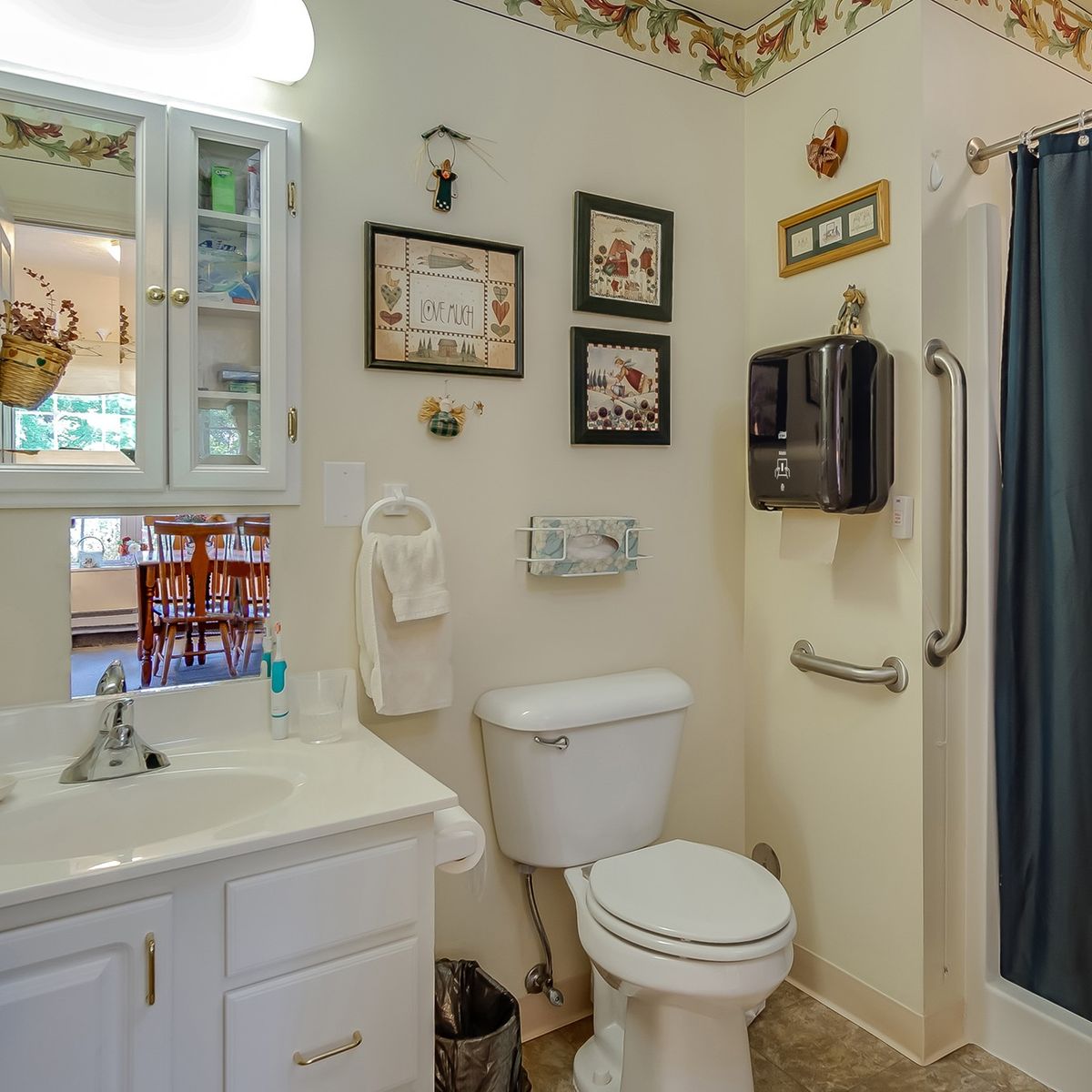 A cozy bathroom with a toilet, sink, and shower curtain, adorned with framed decorations and equipped with a handrail.