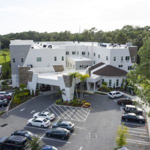 Aerial view of a large white building with an adjacent parking lot surrounded by trees.