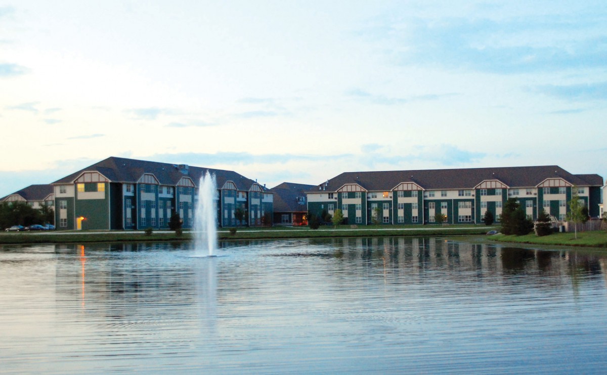 A serene view of buildings by a lake with a central fountain and reflections in the water.