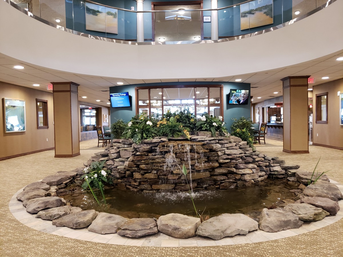 A modern indoor lobby featuring a central stone fountain surrounded by plants.