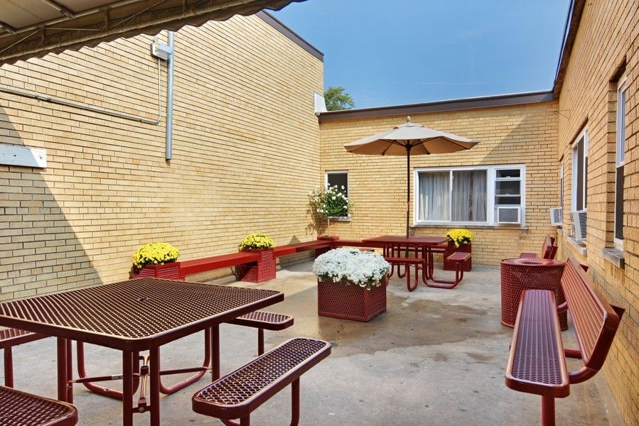 A cozy outdoor patio area with benches, tables, and vibrant flower arrangements against a brick building.