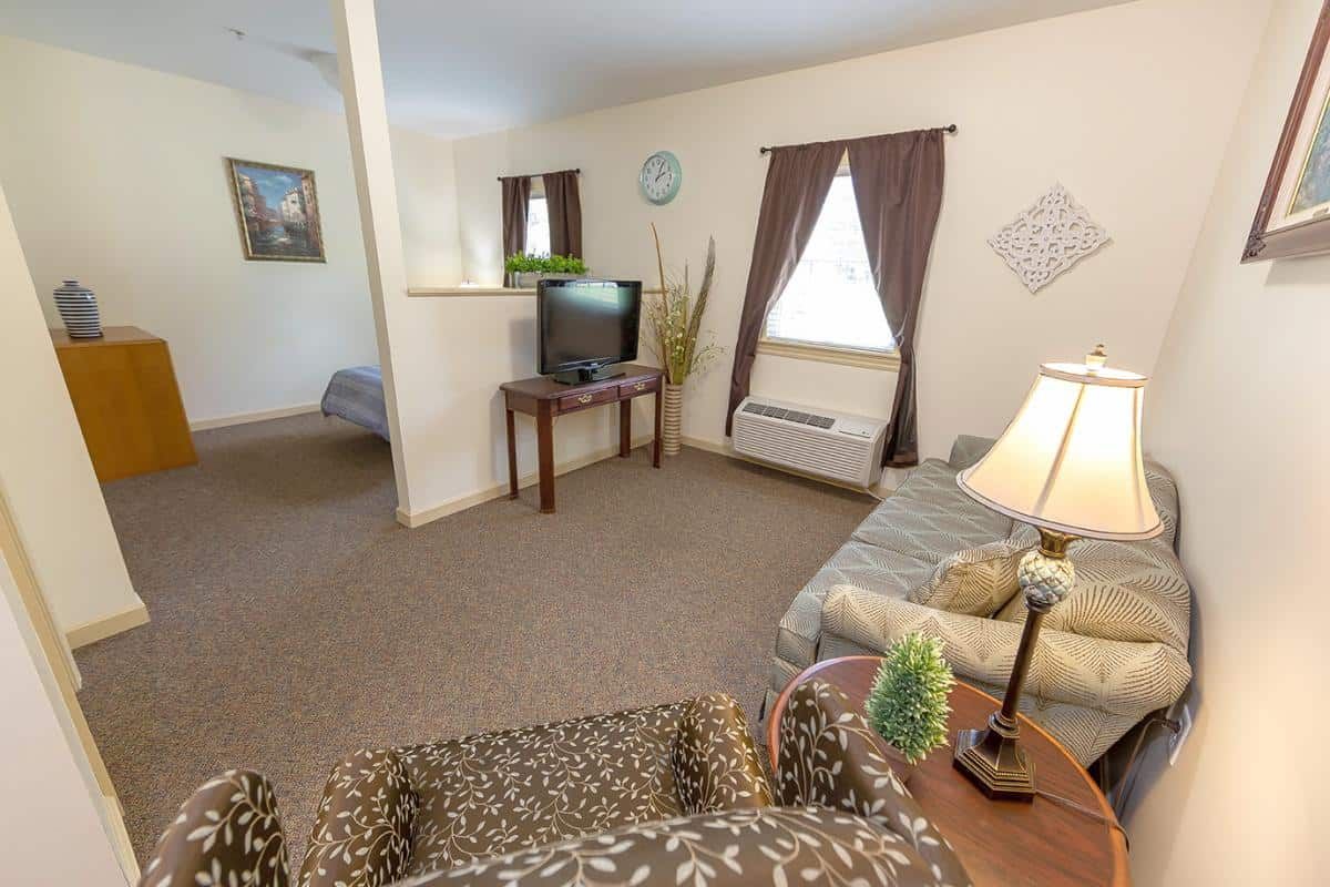 A cozy living room with a sofa, TV, and lamp, featuring brown curtains and a carpeted floor.
