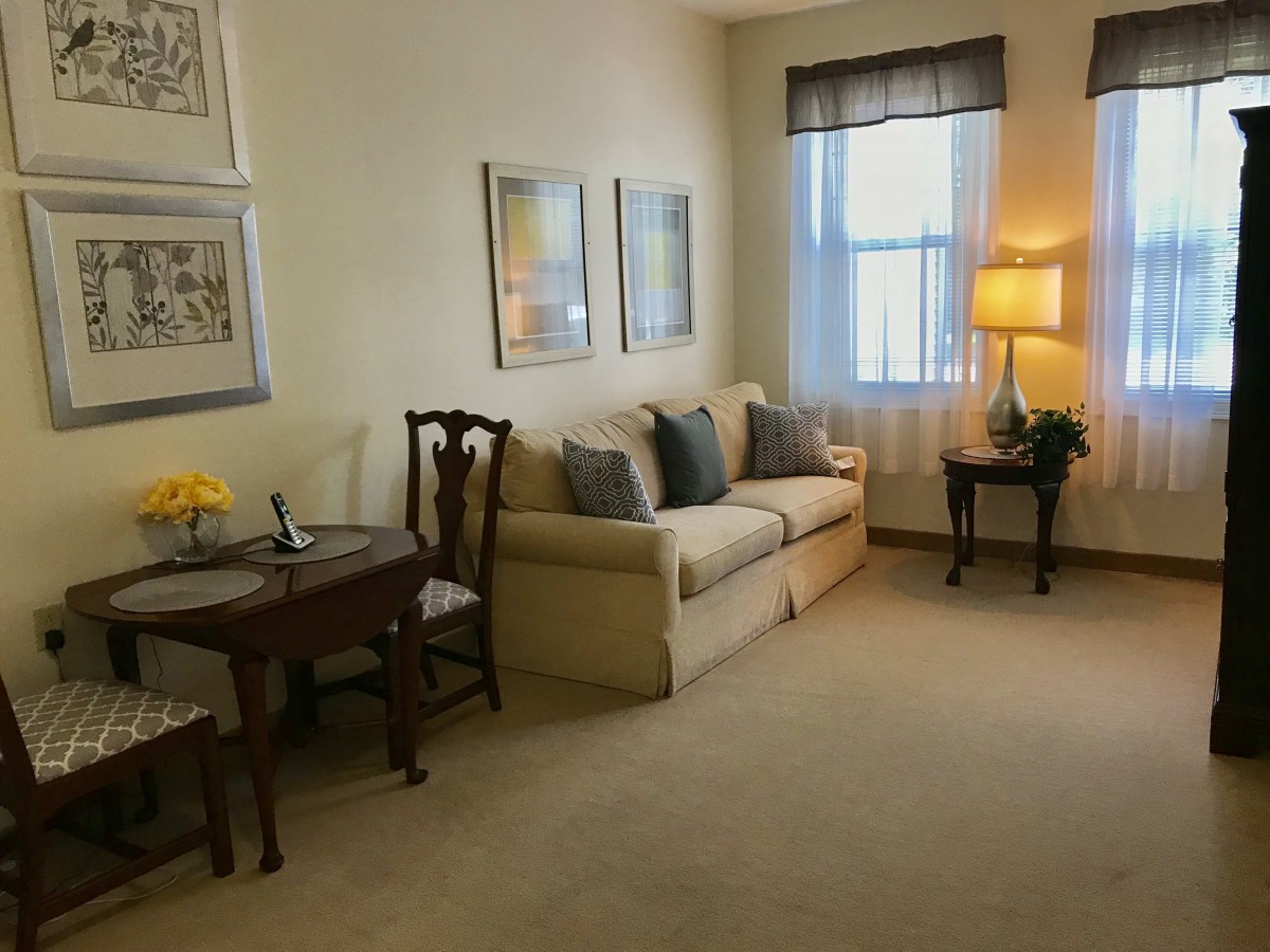 A cozy living room with a sofa, a small table with chairs, and a lamp by the window.