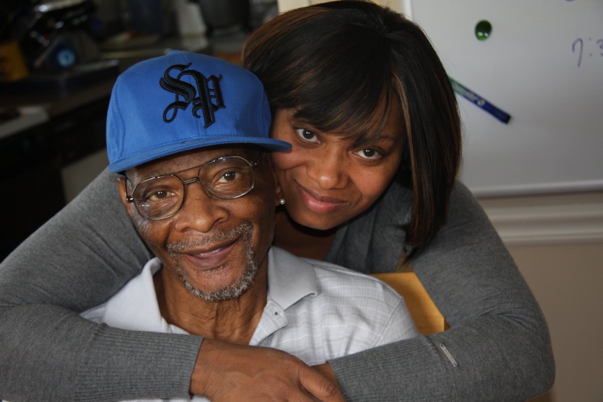 A woman affectionately embraces an elderly man wearing a blue cap and glasses, both smiling warmly.